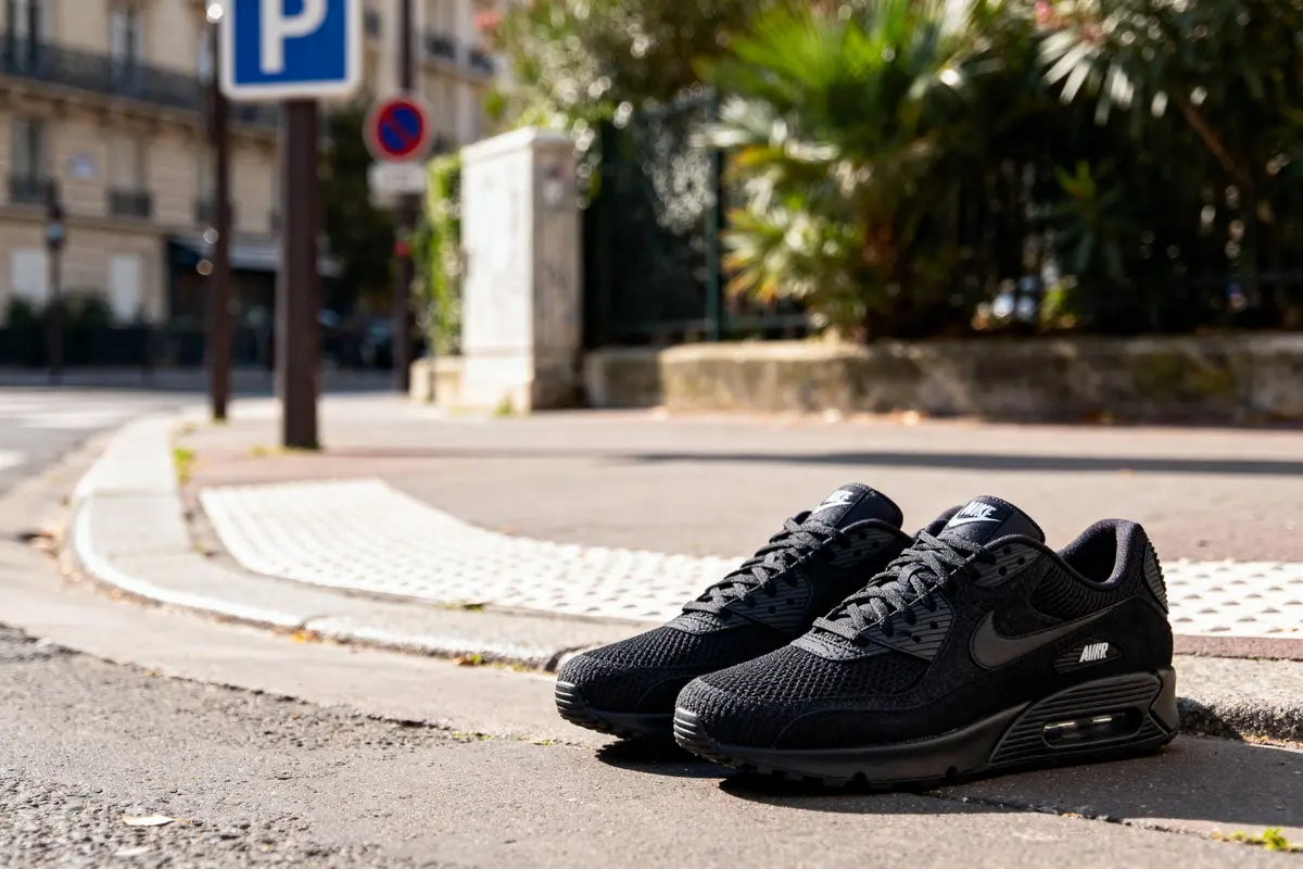 Paire de baskets noires Nike Air Max posées sur un trottoir urbain avec panneaux de signalisation en arrière-plan.