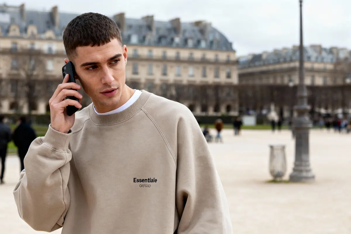 Homme en sweat beige "Essentale" au téléphone, dans un parc urbain avec bâtiments historiques en arrière-plan.