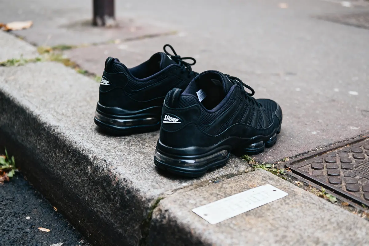 Paire de baskets noires posée sur un trottoir en béton, avec semelles épaisses et étiquette visible.