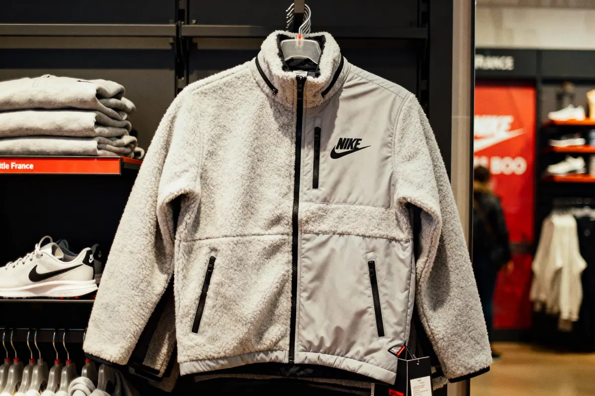 Veste Nike grise en exposition sur cintre, magasin, étagère avec vêtements pliés et chaussures.