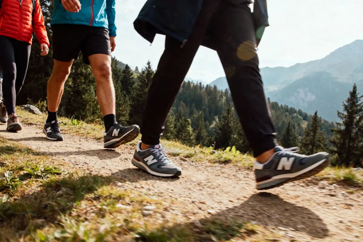 Personnes marchant sur un sentier de randonnée en montagne, portant des baskets New Balance. Paysage boisé en arrière-plan.