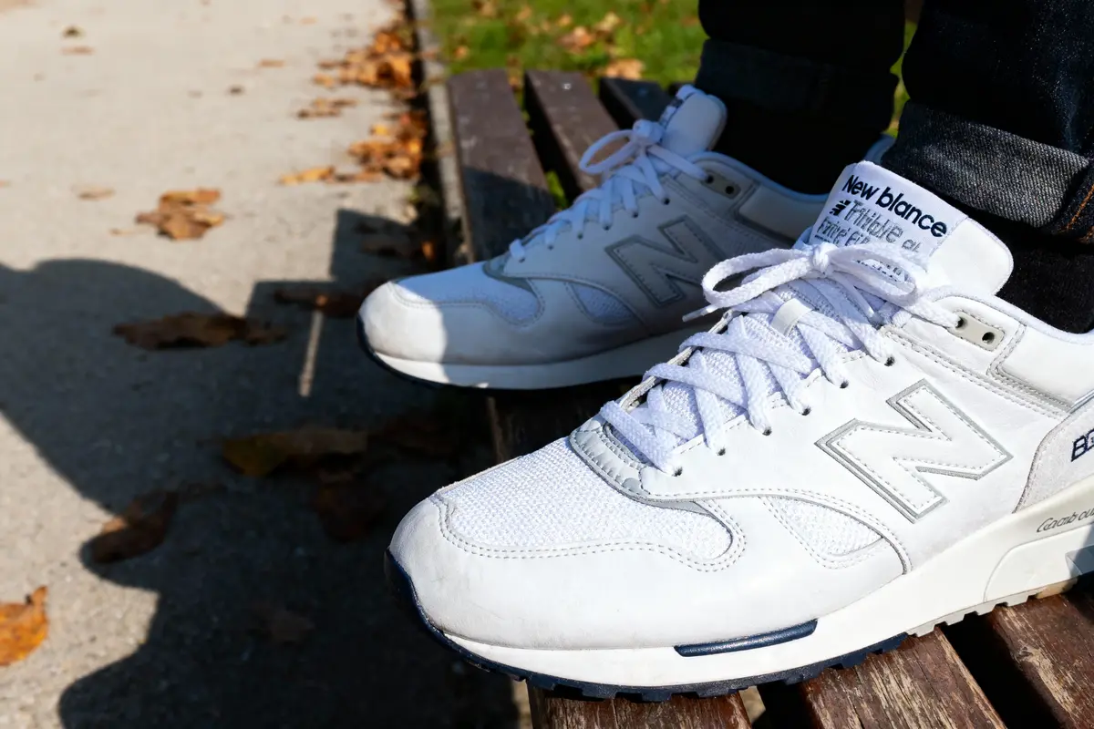 Des baskets blanches New Balance sur un banc en bois, avec feuilles mortes sur le trottoir.
