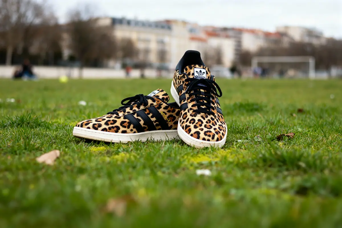 Chaussures léopard Adidas sur une pelouse verte avec bâtiments flous en arrière-plan.