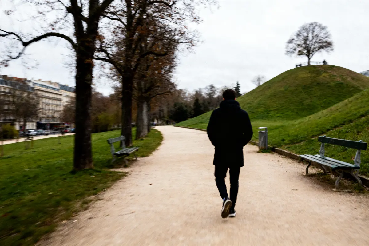 Personne marchant sur un chemin bordé d’arbres et de bancs dans un parc, ciel nuageux.