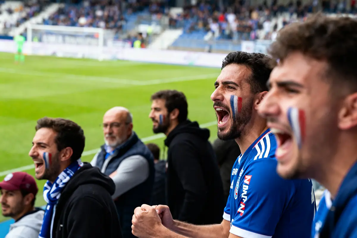 Supporters de football enthousiastes avec peinture faciale, vêtus de bleu, regardant un match depuis les gradins du stade.