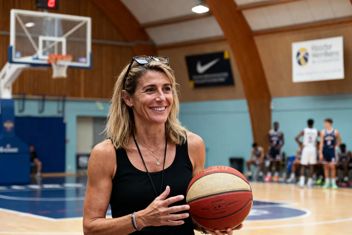 Femme souriante tenant un ballon de basket dans un gymnase, fond flou de joueurs et panier.