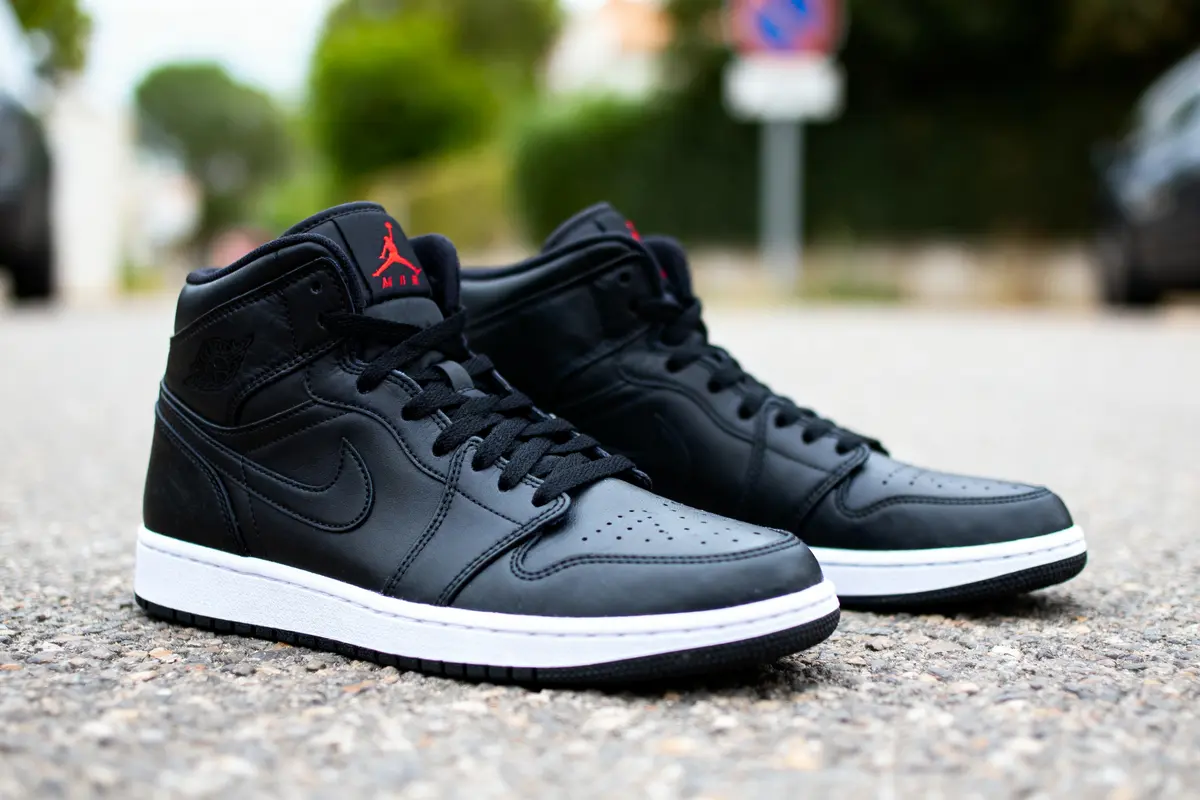 Chaussures noires de basket style urbain posées sur route, arrière-plan flou avec arbres et poteaux.