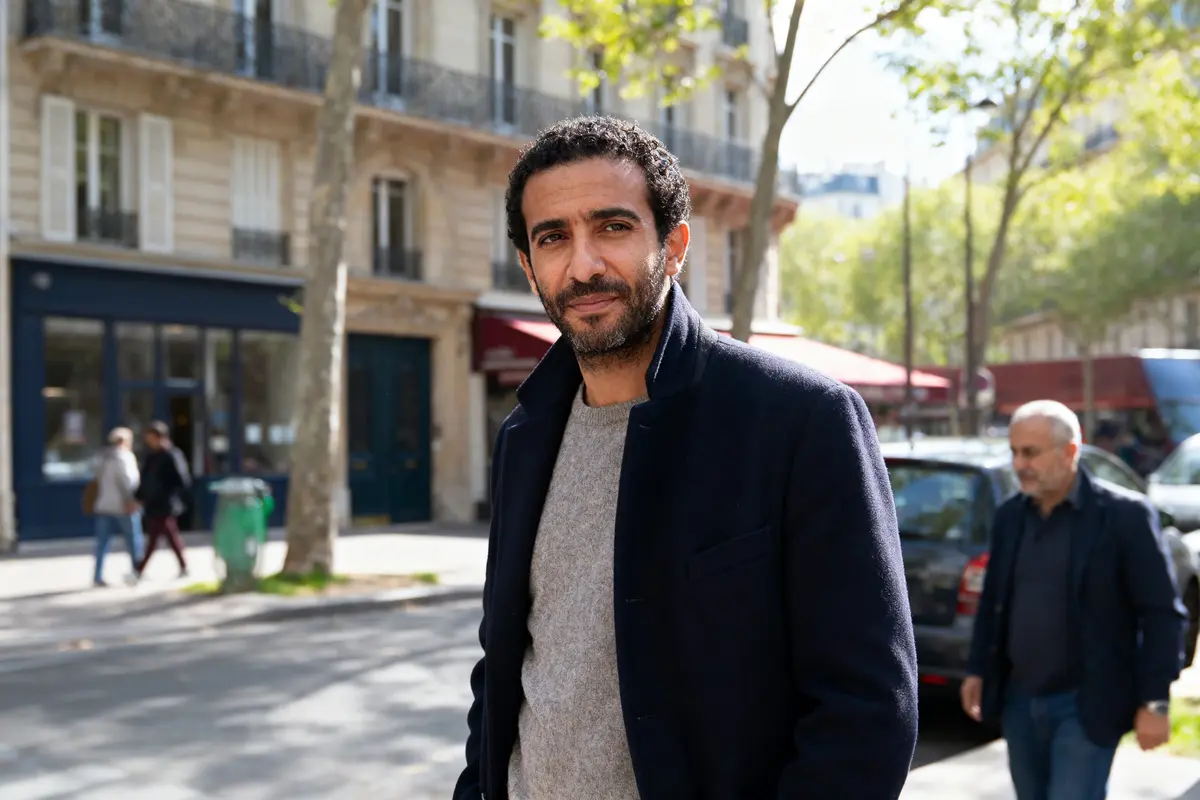 Homme souriant marchant dans une rue parisienne ensoleillée, avec bâtiments et passants en arrière-plan.