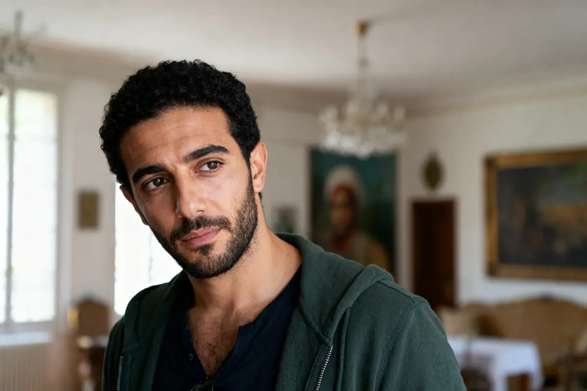 Homme barbu avec sweat à capuche dans pièce lumineuse, peintures floues en arrière-plan.