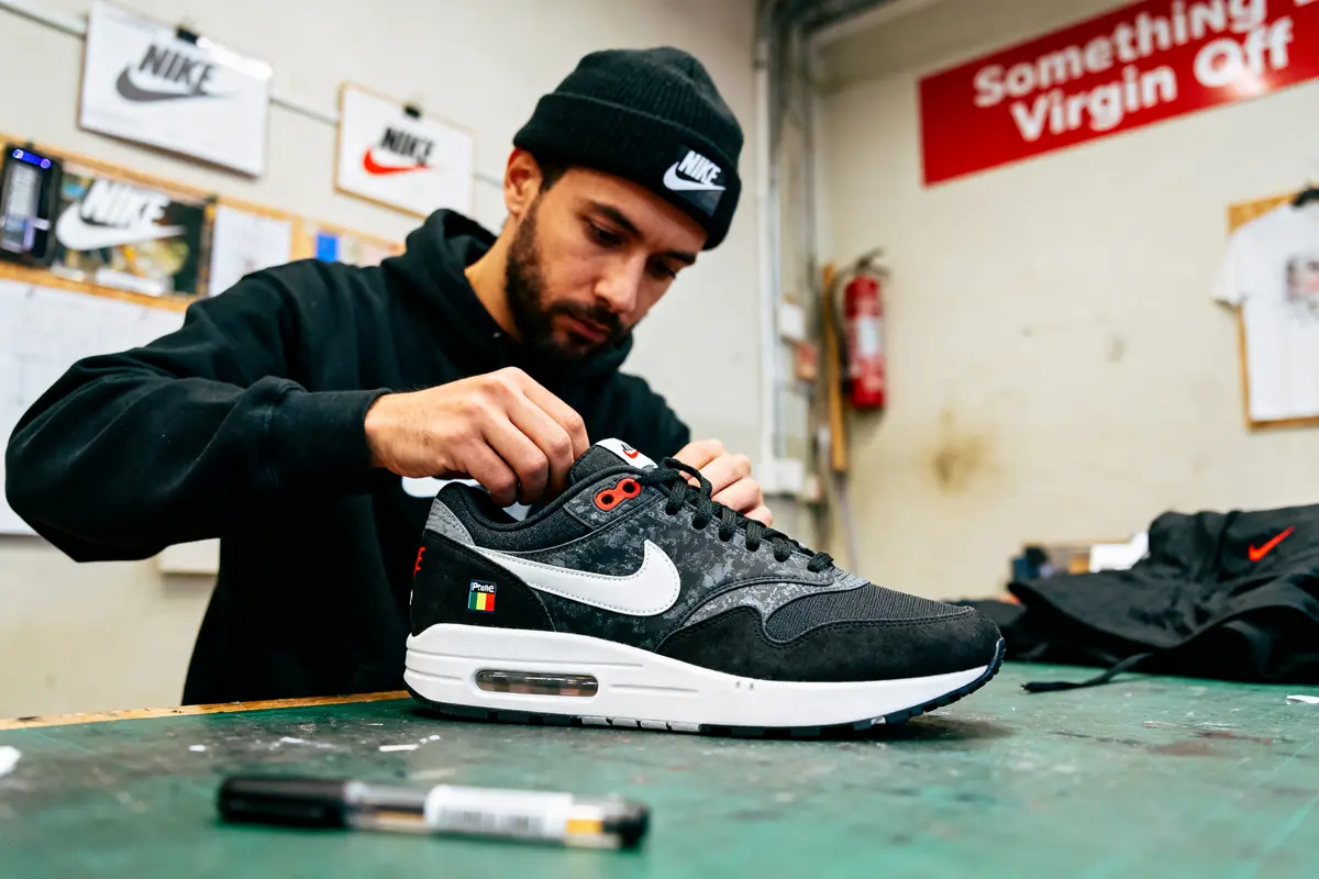 Un homme ajuste une chaussure Nike noire sur une table, portant bonnet noir, arrière-plan atelier.