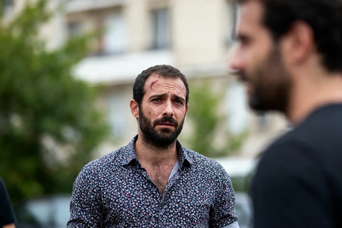 Homme barbu avec chemise à motifs, l'air préoccupé, discutant avec un autre homme en extérieur.