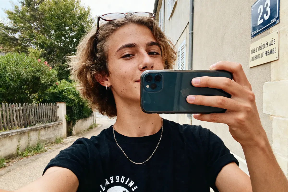 Jeune personne prenant un selfie en extérieur, près d'un bâtiment, portant un t-shirt noir et lunettes sur la tête.