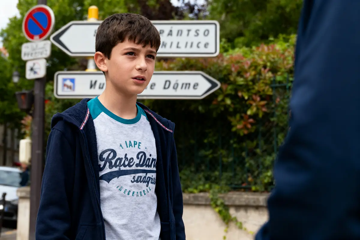 Un garçon brun en t-shirt et veste bleue, devant des panneaux de signalisation dans la rue.