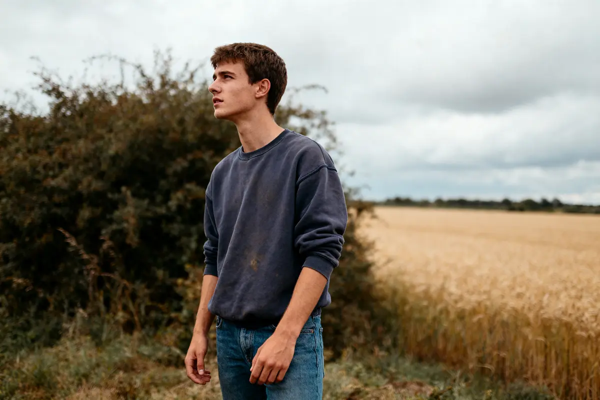 Jeune homme en pull bleu dans un champ de blé sous ciel nuageux.