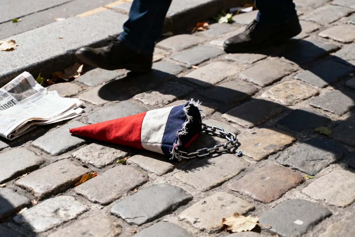 Cône tricolore abîmé et journaux sur pavés, deux personnes passent.