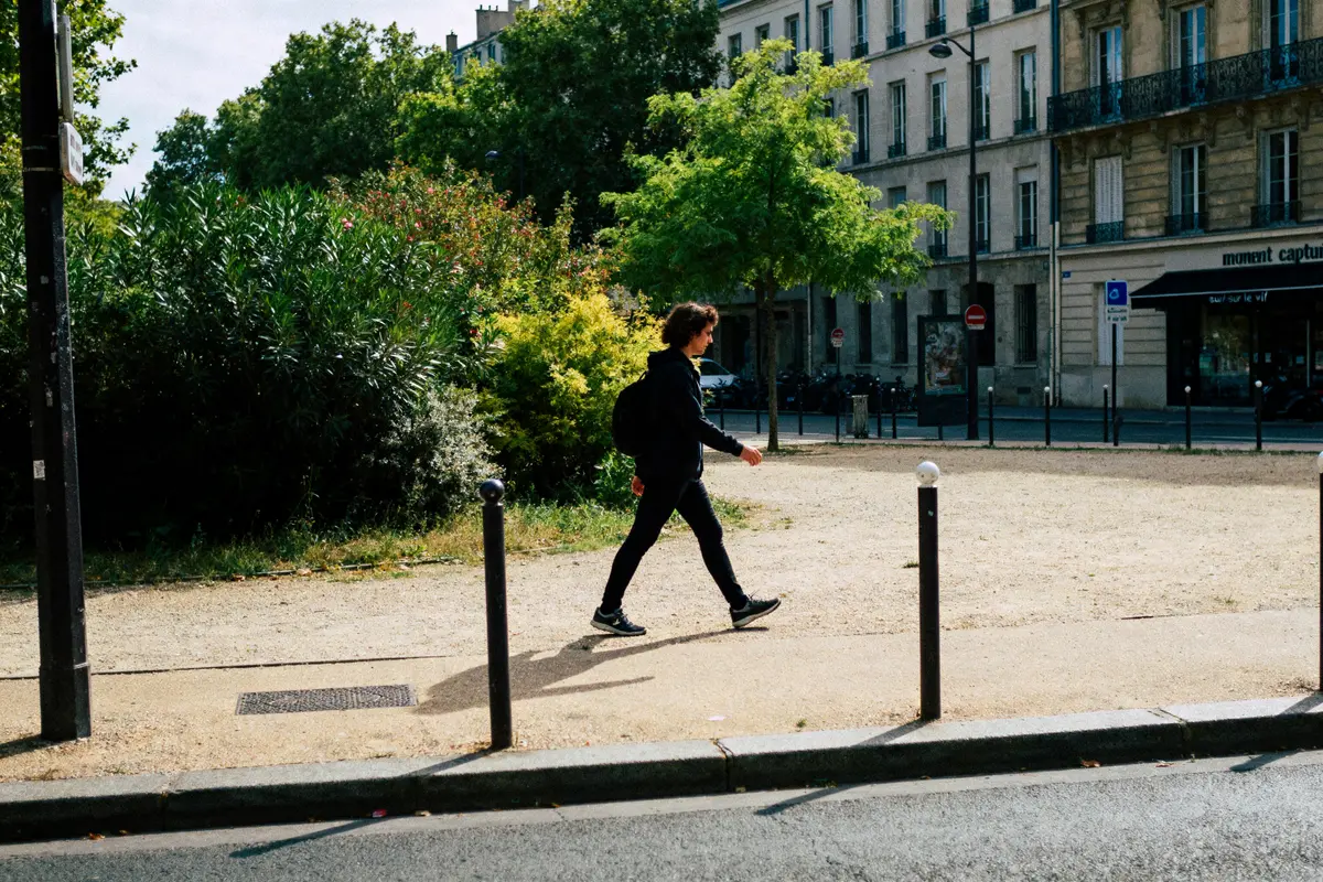 Personne marchant sur un trottoir, avec des arbres et immeubles en arrière-plan, par une journée ensoleillée.