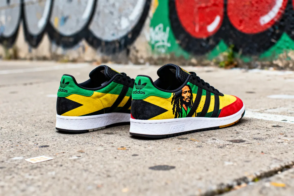 Chaussures Adidas multicolores inspirées de Bob Marley, posées sur un sol devant un mur graffiti.