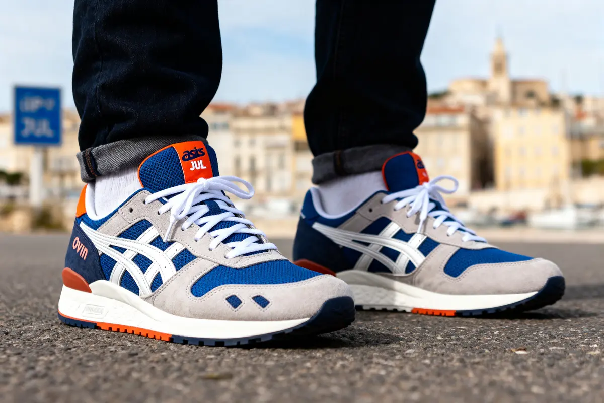 Personne portant des baskets bleues et blanches Asics sur fond urbain flou, chaussée en premier plan.
