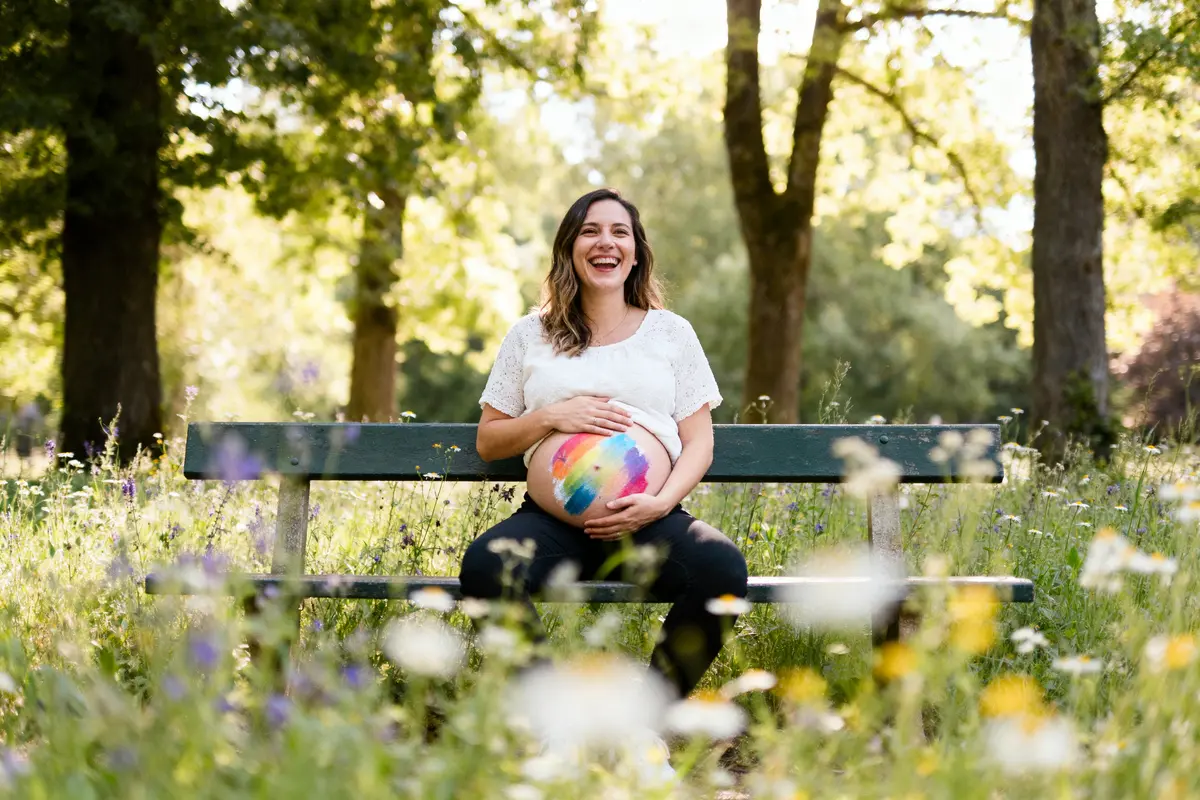 Une femme enceinte assise sur un banc, tient son ventre peint de couleurs vives, souriante, parc verdoyant.