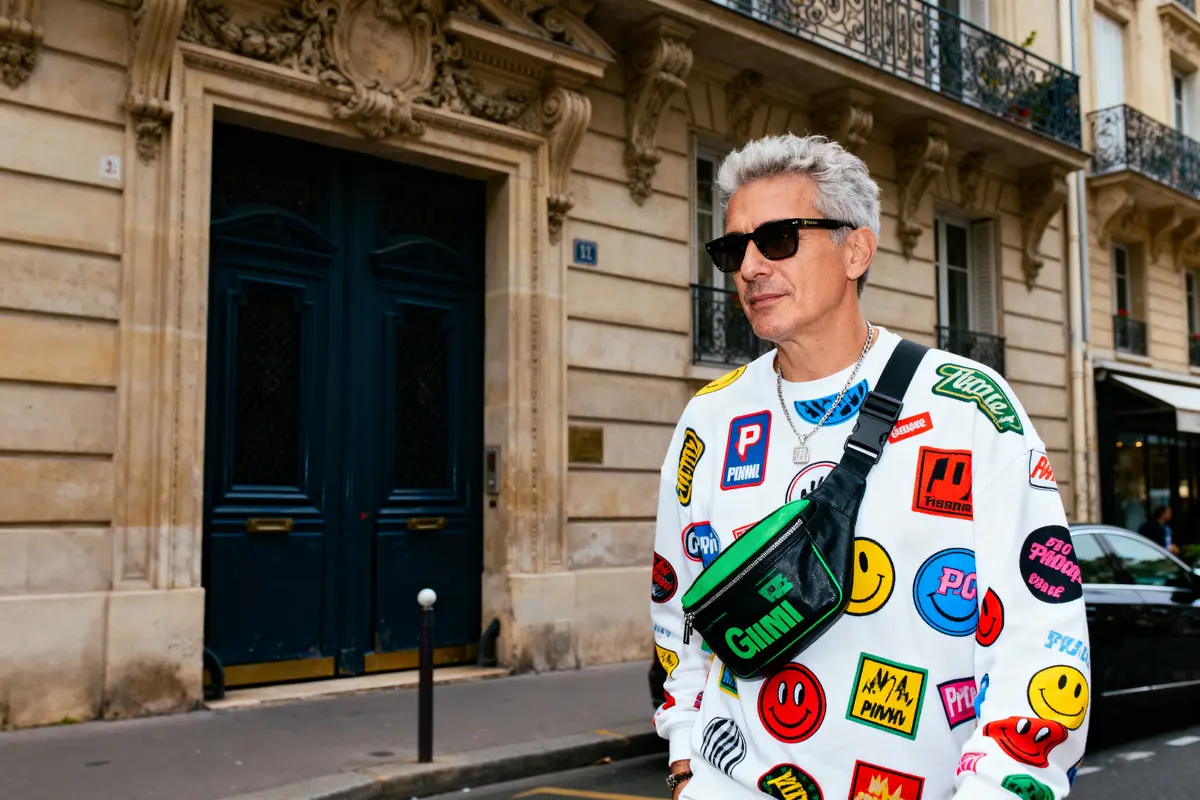 Homme portant un pull coloré avec logos, sacoche noire, lunettes de soleil, devant un bâtiment parisien élégant.
