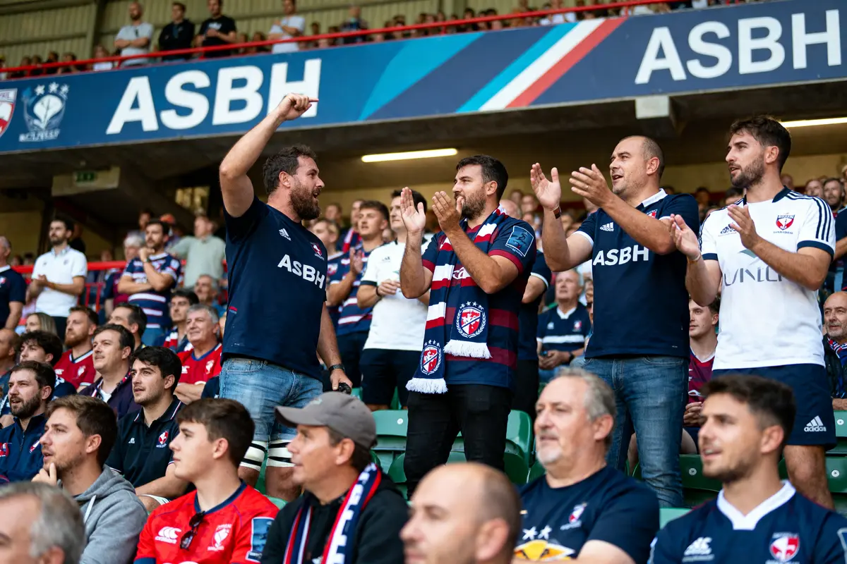 Des supporters en tenue ASBH applaudissent et chantent dans les gradins d'un stade.
