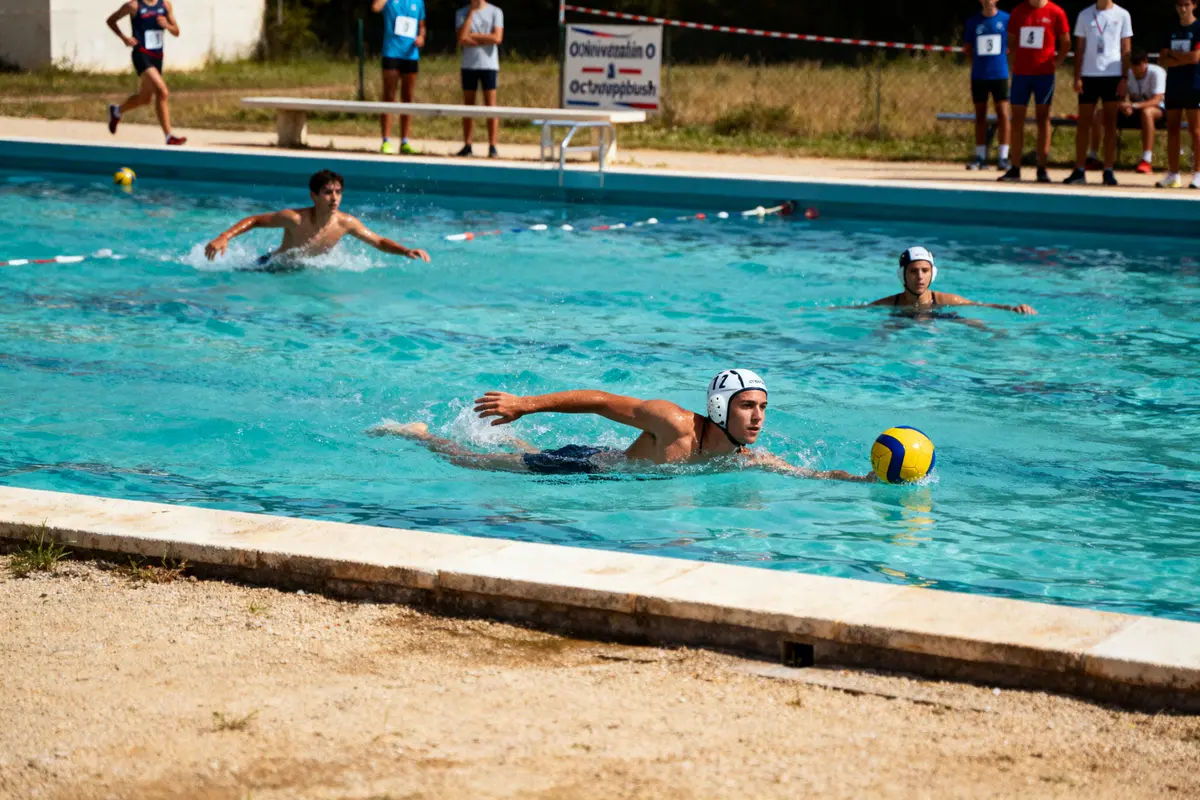 Joueurs de water-polo dans une piscine extérieure, participant à un match ou à un entraînement, spectateurs en arrière-plan.