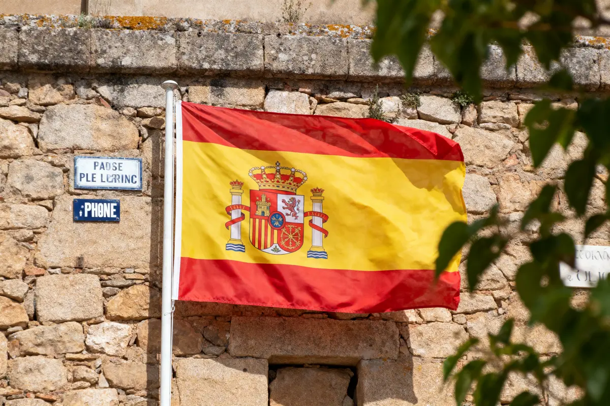Drapeau espagnol flottant sur un mur de pierre, panneau de rue et branches floues au premier plan.