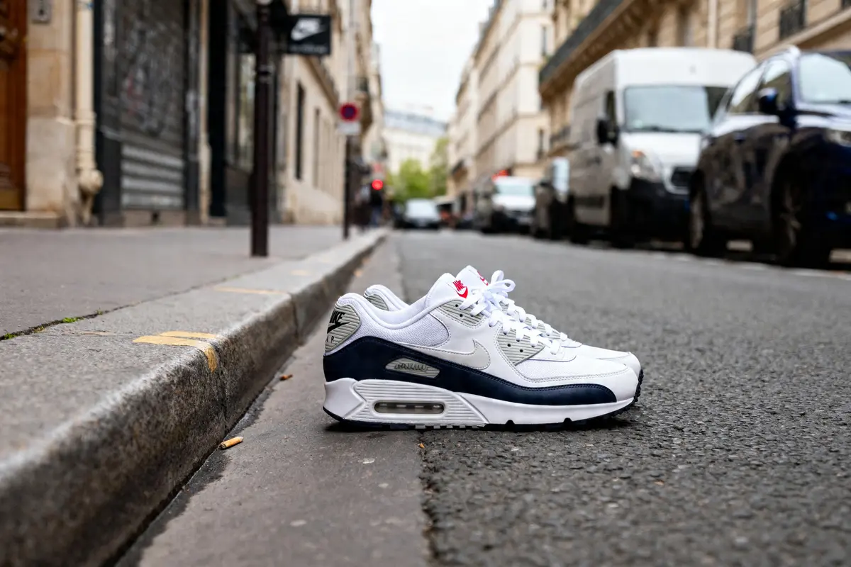 Baskets Air Max 90 blanches sur rue pavée urbaine avec immeubles et voitures en arrière-plan