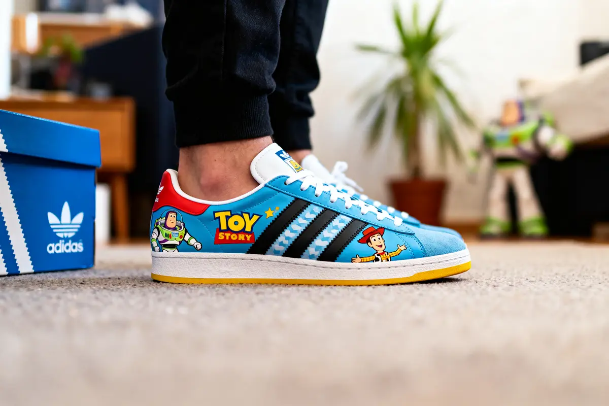 Chaussures Adidas Toy Story bleues avec illustrations Buzz et Woody, boîte bleue, tapis beige, plante en arrière-plan.