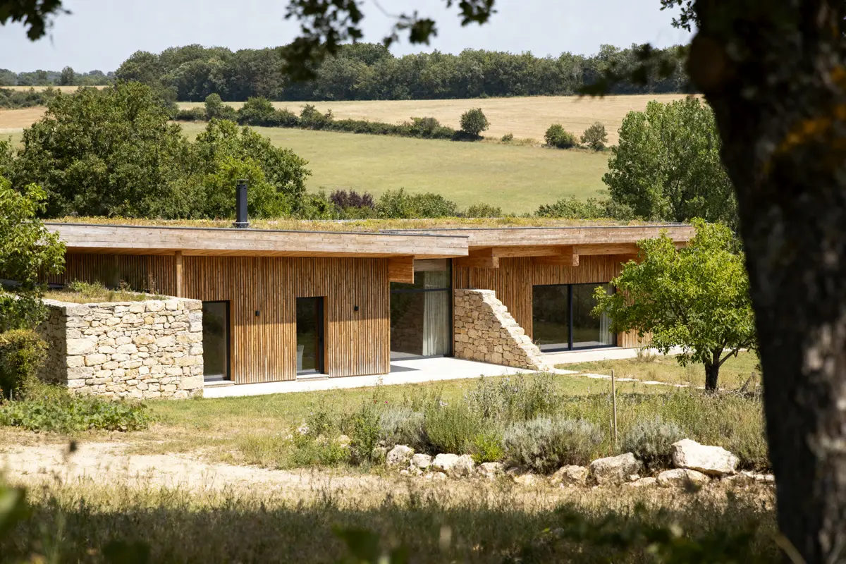 Maison moderne en bois et pierre dans paysage campagnard verdoyant, entourée de champs et arbres.