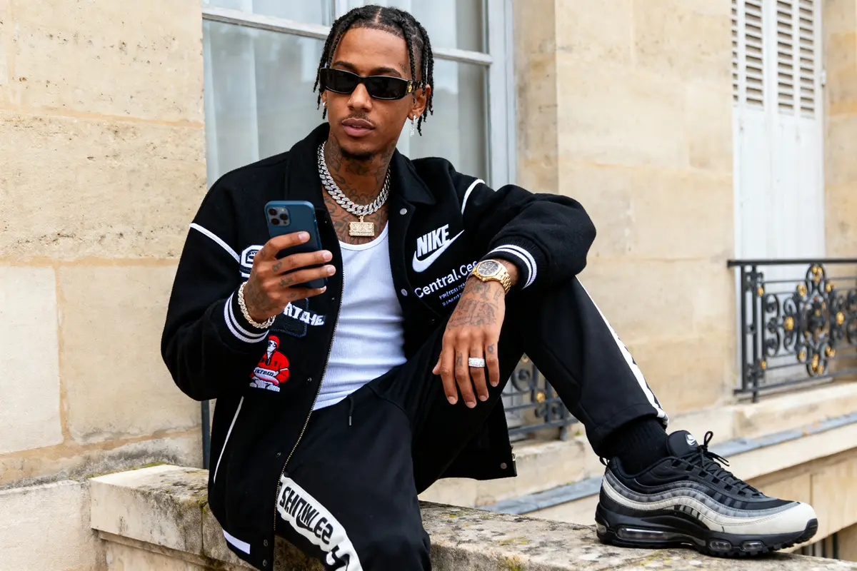Homme assis, portant vêtements de sport, regardant son téléphone, lunettes de soleil et bijoux, devant bâtiment en pierre.