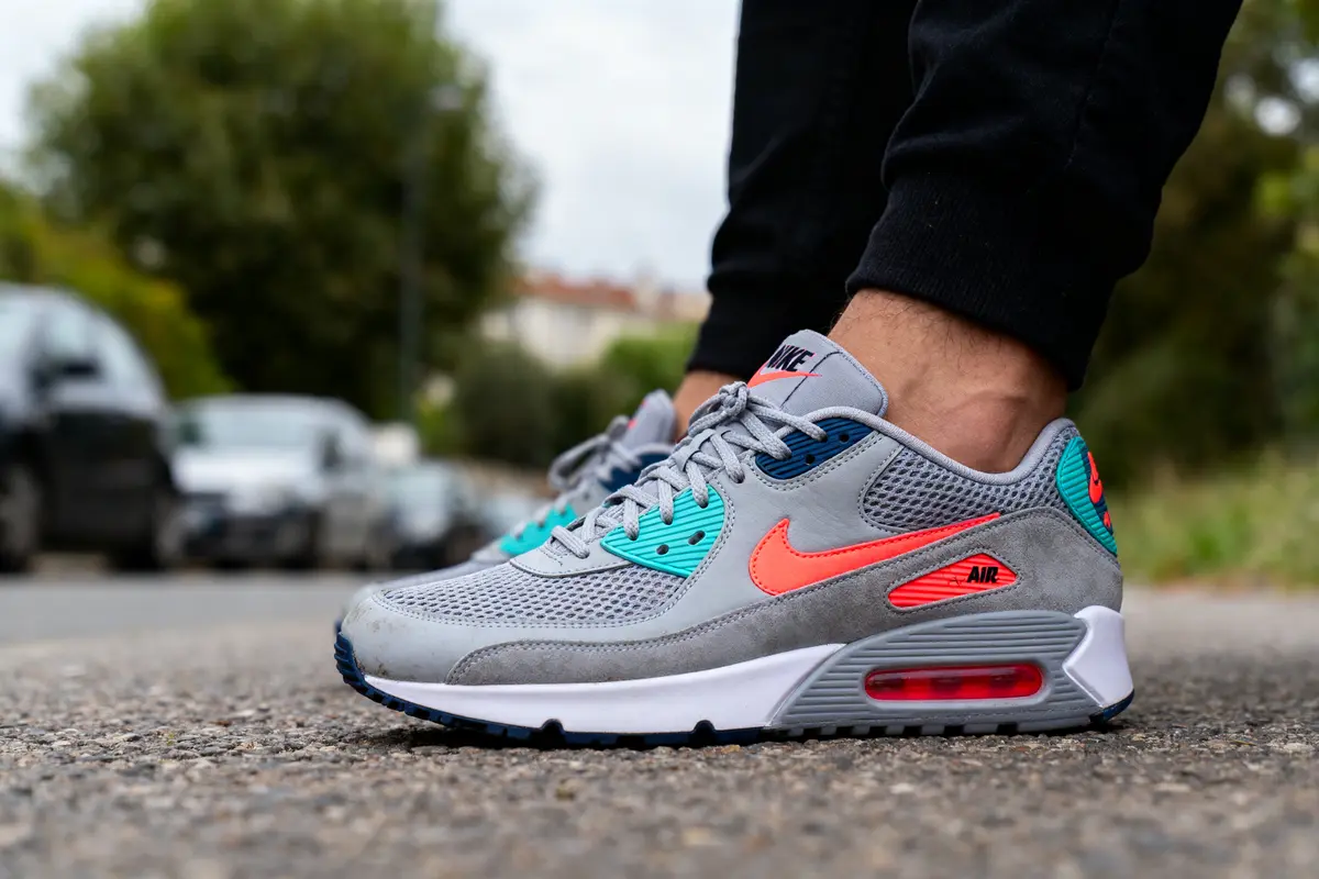 Baskets Nike grises avec détails orange et turquoise, portées par une personne sur un trottoir.