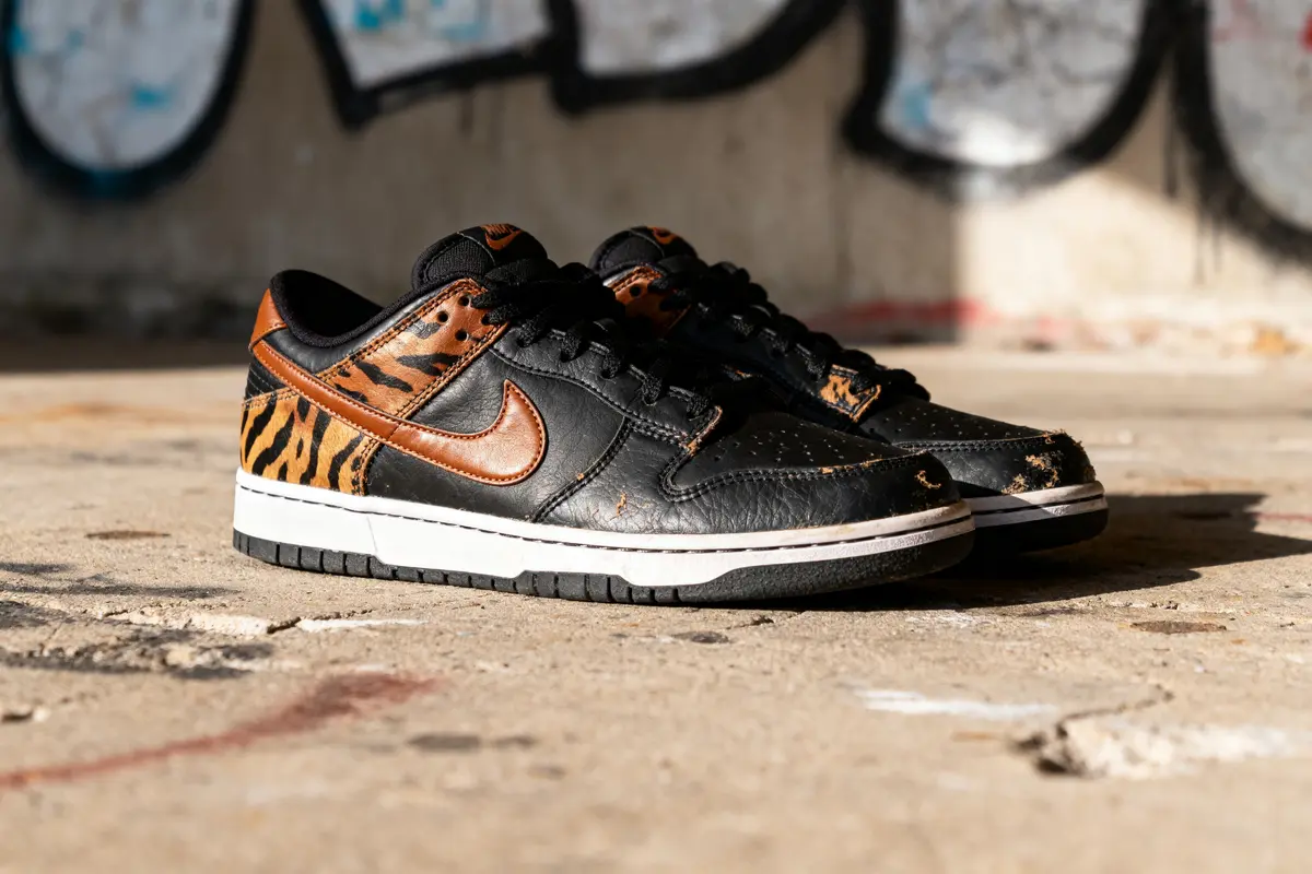 Chaussures noires avec imprimé tigre, posées sur sol en béton avec graffiti en arrière-plan.