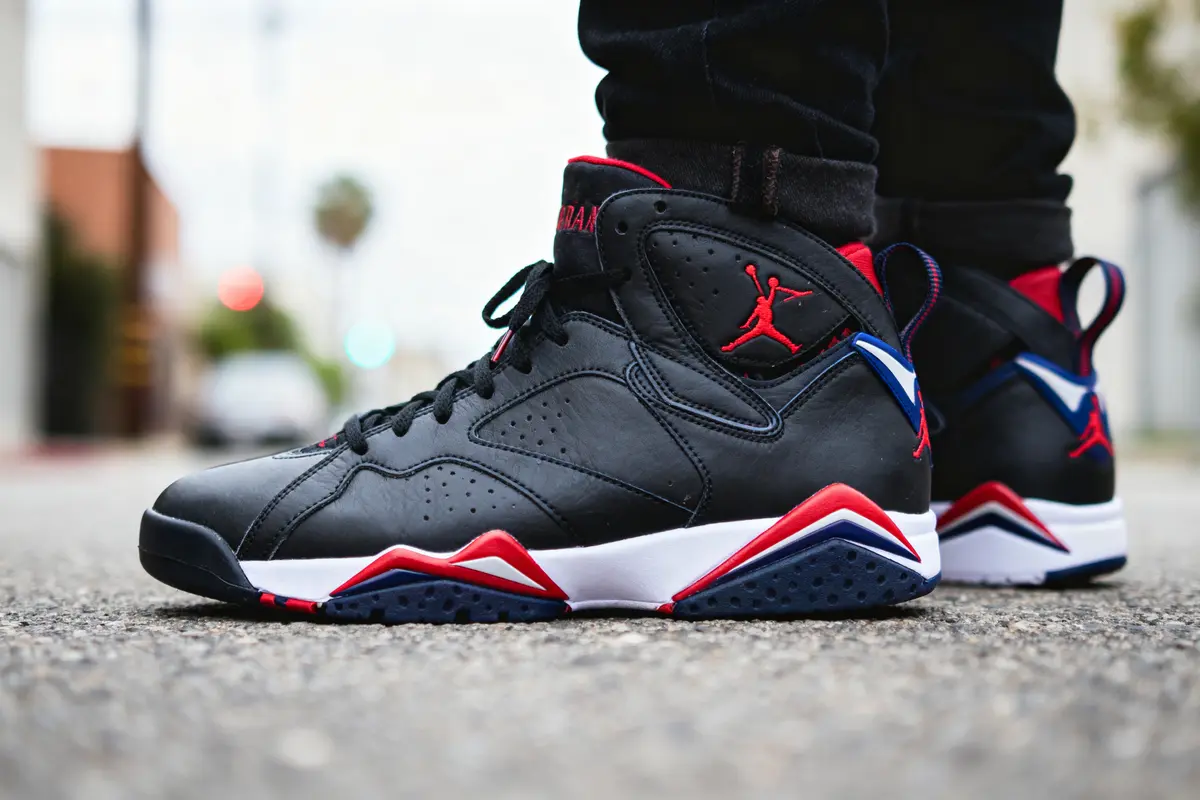 Chaussures de sport noires, rouges et blanches sur sol urbain, personne debout en arrière-plan flou.