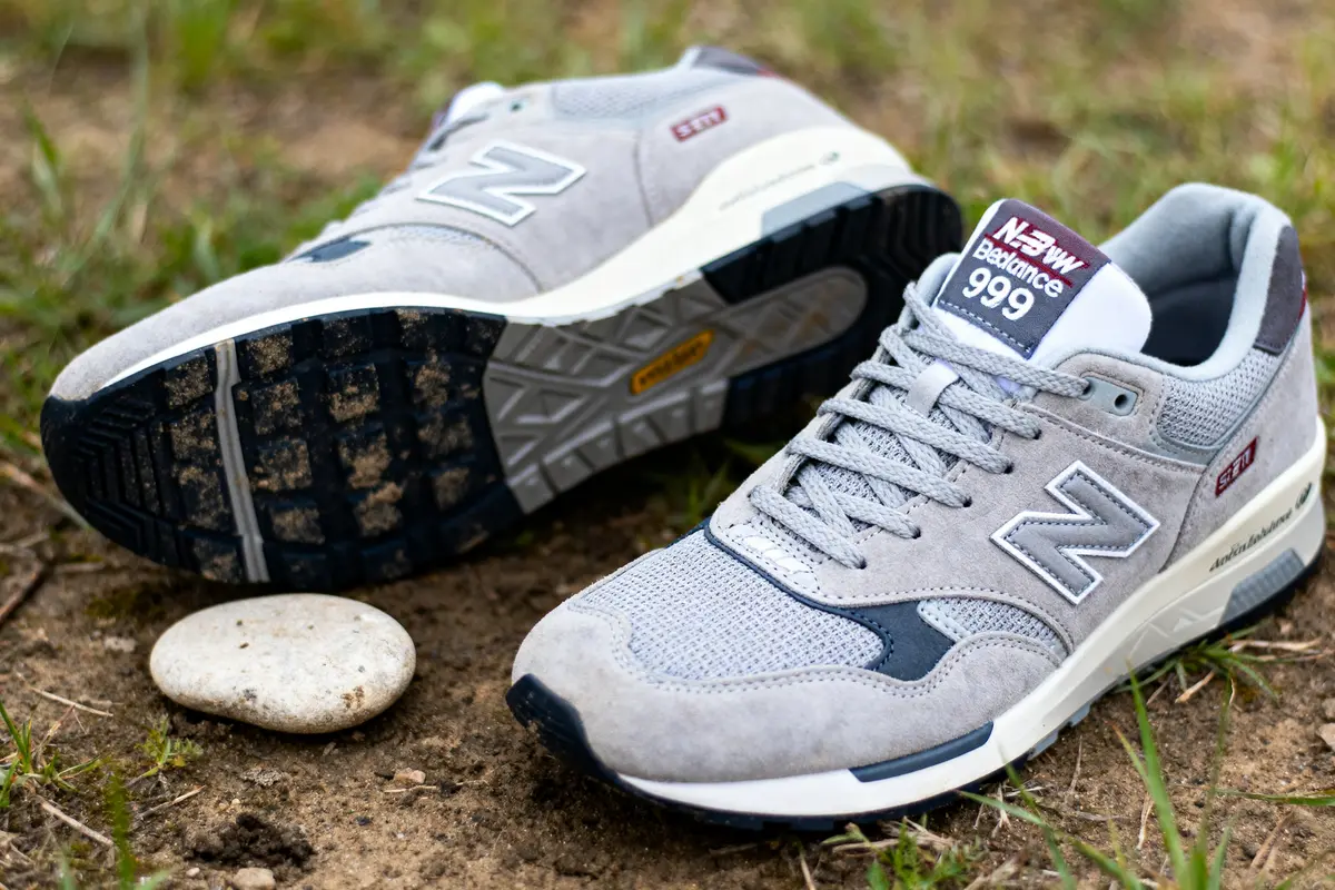 Chaussures de sport grises New Balance 999, posées sur sol herbeux et terreux avec un caillou.
