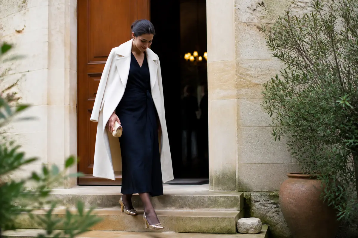 Femme élégante descendant des marches, portant un manteau crème, une robe noire et des talons dorés.